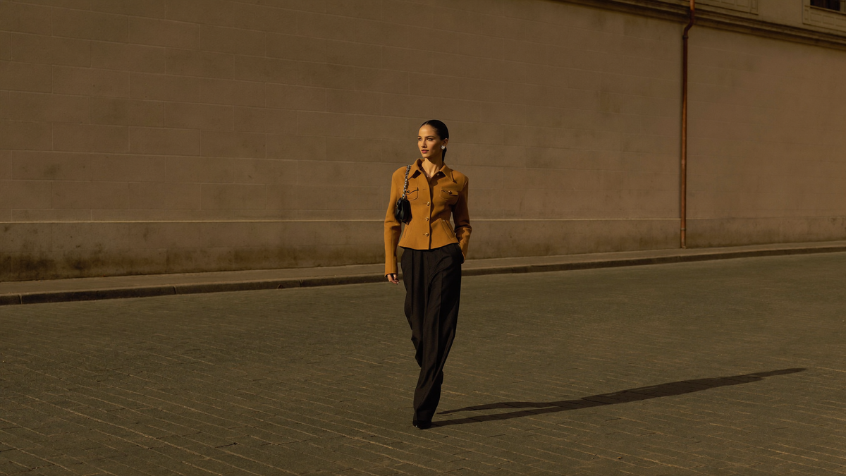 Woman in a tan jacket walking down a sunlit street